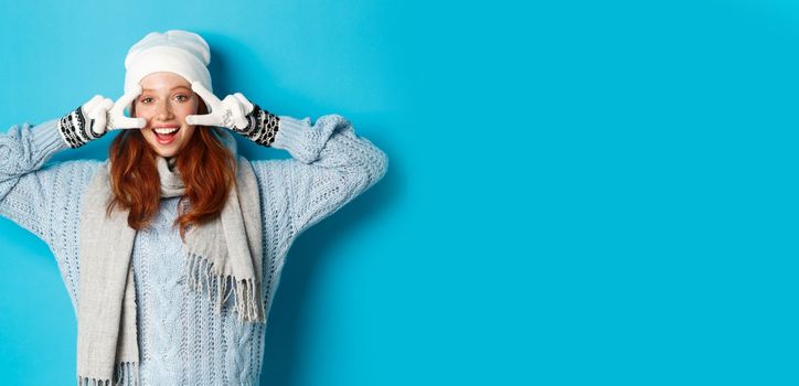 Winter and holidays concept. Cute redhead teen girl in beania, gloves and sweater showing peace sign, looking left at camera and wishing merry christmas, standing against blue background.