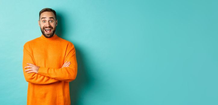 Image of happy and surprised man reacting to news, looking amazed, standing in orange sweater against turquoise background.