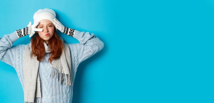 Winter and holidays concept. Happy redhead girl in beanie hat, scarf and gloves showing peace sign and pucker lips sassy, staring at camera, wearing sweater, posing over blue background.