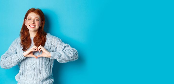 Cute redhead girl in sweater showing heart sign, I love you gesture, smiling at camera, standing against blue background.