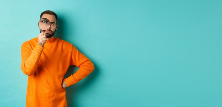 Funny man looking serious through magnifying glass, inspecting something, standing in orange sweater against turquoise background.