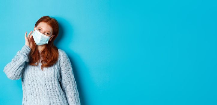 Winter, covid-19 and social distancing concept. Cute redhead teenage girl, wearing face mask and tuck hair strand behind ear, staring at camera, standing over blue background.