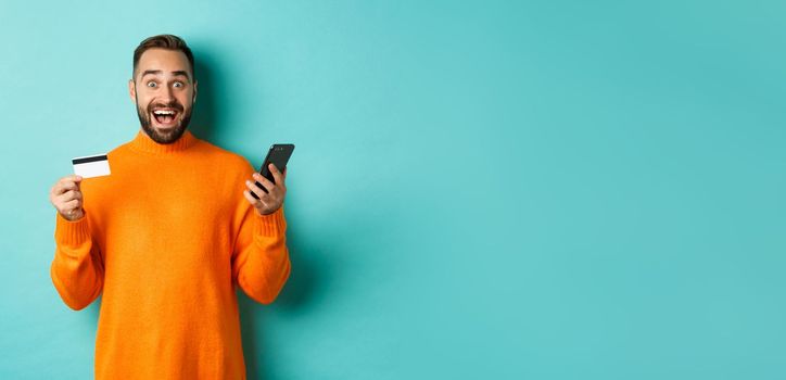 Online shopping. Surprised man holding mobile phone and credit card, paying in internet store, standing over light blue background.