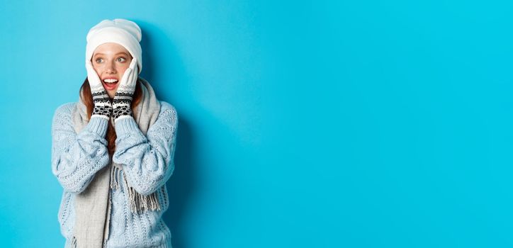 Excited redhead girl staring left at logo, wearing winter clothes, beanie, gloves and sweater, standing over blue background.