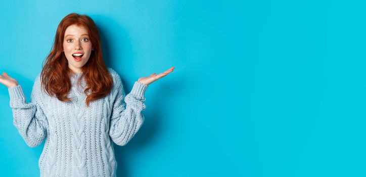 Image of redhead young woman spread hands sidewas, looking with disbelief and amazement, hear news, standing over blue background.