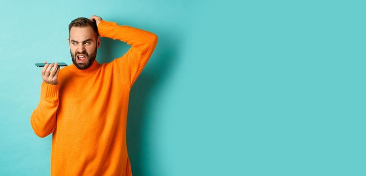 Confused man scratching head while talking on speakerphone, record voice message with indecisive face, standing in orange sweater over light blue background.