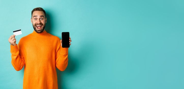 Online shopping. Excited man showing mobile screen and credit card, smiling amazed, standing over turquoise background.