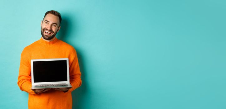 Happy caucasian man thinking and showing laptop screen, smiling pleased while pondering, standing in orange sweater against light blue background.