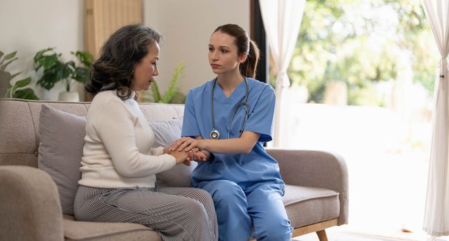 Happy patient is holding caregiver for a hand while spending time together. Elderly woman in nursing home and nurse...