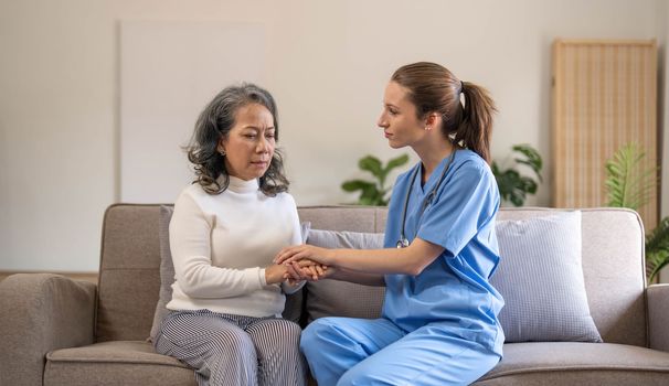 Happy patient is holding caregiver for a hand while spending time together. Elderly woman in nursing home and nurse...