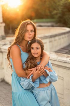 Portrait of mother and daughter in blue dresses with flowing long hair against the backdrop of sunset. The woman hugs and presses the girl to her. They are looking at the camera