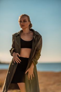 Portrait blonde sea cape. A calm young blonde in an unbuttoned khaki raincoat stands on the seashore, under the raincoat there is a black skirt and top.