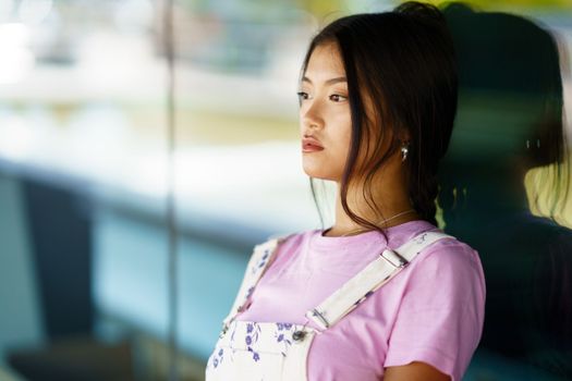 Stylish young Asian woman with black hair leaning on glass wall and looking away in daytime on city street