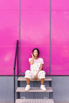 Full body young Asian female in stylish clothes touching hair and looking at camera while sitting on stairs near pink wall