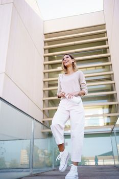 Low angle of cheerful female in stylish outfit, laughing and looking away while standing on path and opening eco friendly bottle of water against modern building on city street