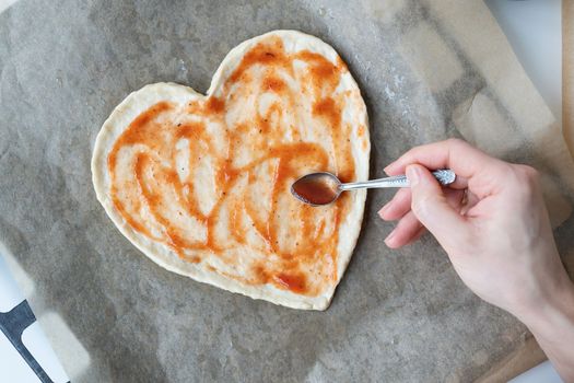 Pizza dough in the shape of a heart, the chef spreads tomato sauce on the pizza. The concept of a surprise for St. Valentine's Day