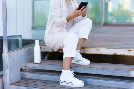 Unrecognizable woman taken a coffee break, sitting on steps near her office building with an eco-friendly ecological metal water bottle. Businesswoman using smartphone.