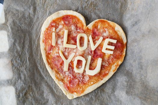 Italian heart shaped pizza with salami, tomato sauce, parmesan, pizza sauce, mozzarella and olive oil on parchment paper with cheese I love you. Love concept for Valentine's Day February 14