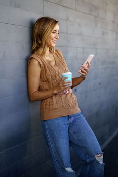 Happy adult woman with coffee to go smiling and watching video on cellphone while leaning on gray wall on city street