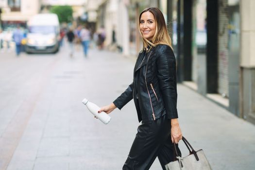 Side view of stylish female in leather pants and jacket carrying bag and takeaway reusable bottle while commuting on town street looking at camera