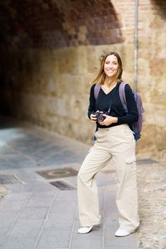 Confident happy young female tourist with long brown hair in casual outfit, and backpack smiling brightly and looking at camera while standing on old town with photo camera