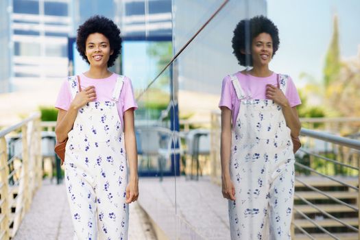 Happy African American young female with Afro hairstyle in overall looking away with smile while standing near glass building in city