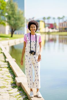 Full body of glad African American female with photo camera looking away while standing near lake on sunny summer day