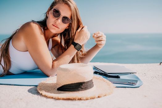 Successful business woman in yellow hat working on laptop by the sea. Pretty lady typing on computer at summer day outdoors. Freelance, travel and holidays concept.