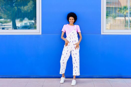 Full body of positive African American female in overall looking at camera while standing near blue house on paved street