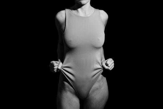 Black and white of crop unrecognizable fit alluring female in bodysuit standing in dark studio with clenched fists