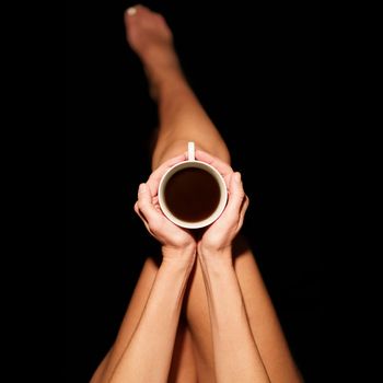 Top view of crop unrecognizable barefooted female sitting in black studio with cup of hot coffee