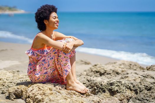 Cheerful young black female traveler in summer dress, with curly hair embracing knees and smiling with closed eyes while sitting on uneven stones against waving sea on sunny day on beach