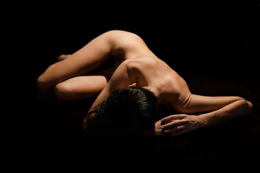 Full body of anonymous fit naked female with short dark hair lying on floor against black background in studio