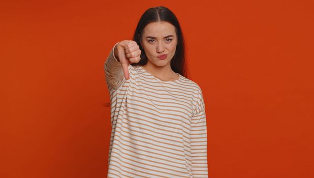 Dislike. Upset unhappy pretty woman in pullover showing thumbs down sign gesture, expressing discontent, disapproval, dissatisfied, dislike. Young adult girl. Indoor studio shot on red background