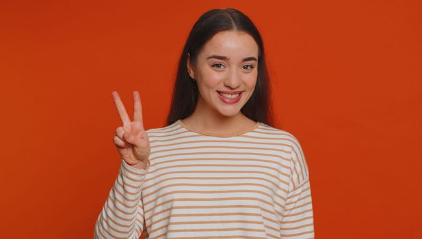 Hipster pretty woman in pullover showing victory v sign, hoping for success and win, doing peace gesture, smiling with kind optimistic expression. Young adult girl isolated on red studio background