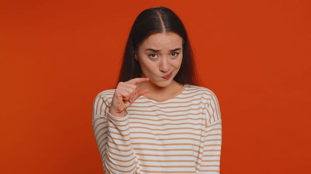 Need some more, please give me. Lovely woman showing a little bit gesture with sceptic smile, showing minimum sign, measuring small size. Young adult girl isolated alone on red studio background