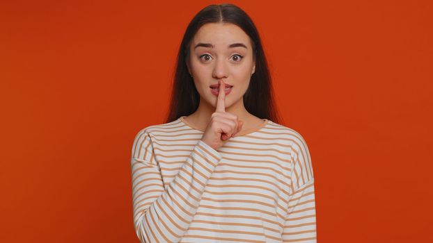 Shh be quiet please. Portrait of millennial woman 20 years old presses index finger to lips makes silence gesture sign do not tells secret. Young lovely pretty girl posing on red studio background