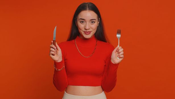 Ready to eat. Hungry young woman in crop top waiting for serving dinner dishes with cutlery, will appreciate delicious restaurant meal menu. Girl holding fork and knife on red studio background