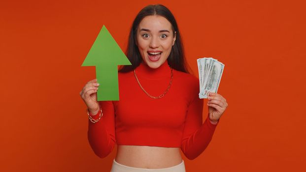 Happy pretty woman winner holding arrow sign pointing up, career growth and money dollar exchange increase. Young adult girl showing up indicating successful development progress on red background