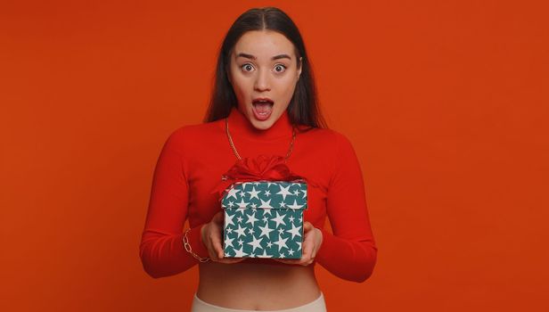 Happy pretty woman opening gift box, unwrapping birthday surprise and expressing great happiness amazement, satisfied with best present bonus. Young adult lovely girl isolated on red studio background