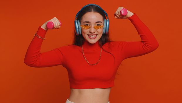 Young fit woman listening music via headphones, working out, pumping up arm muscles lifting pink dumbbells, practicing pilates, healthcare. Pretty girl indoors isolated alone on studio red background