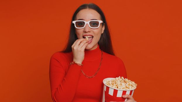 Excited pretty woman in 3D glasses eating popcorn and watching interesting TV home serial, sport game, film, online social media movie content. Girl enjoying domestic entertainment on red background