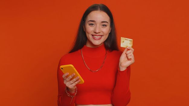 Sincere pretty woman customer using credit bank card and smartphone while transferring money, purchases online shopping, payment. Finance and internet. Young girl isolated alone on red background