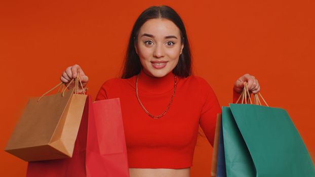 Young woman in crop top showing shopping bags, advertising discounts, smiling looking amazed with low prices, shopping on Black Friday holidays. Girl indoors isolated alone on studio red background