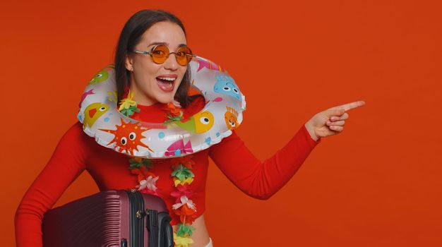 Cheerful happy tourist woman with luggage bag, swimming ring pointing empty place, advertising area for commercial text, copy space for goods services banner promotion. Vacation, summer trip to sea