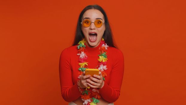 Cheerful tourist woman with hawaiian lei in sunglasses using smartphone celebrating winning holiday resort vacation tickets pass. Tourism, summer trip to sea. Happy pretty girl on red wall background