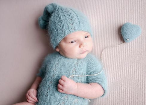 Cute newborn baby boy wearing knitted costume anf hat holding heart toy and looking back. Adorable infant child kid studio portrait