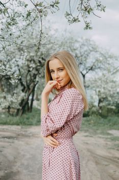 Blonde girl on a spring walk in the garden with cherry blossoms. Female portrait, close-up. A girl in a pink polka dot dress