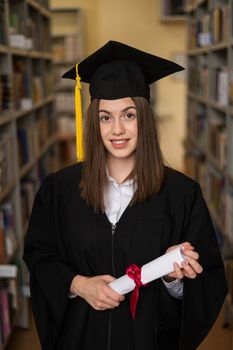 Happy young woman in graduate gown holding diploma in the library