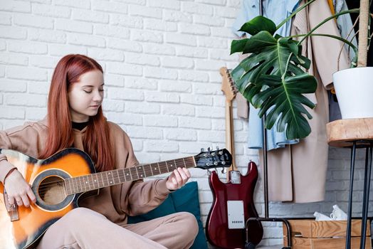 young caucasian red-haired woman sitting on rug and playing acoustic guitar at home